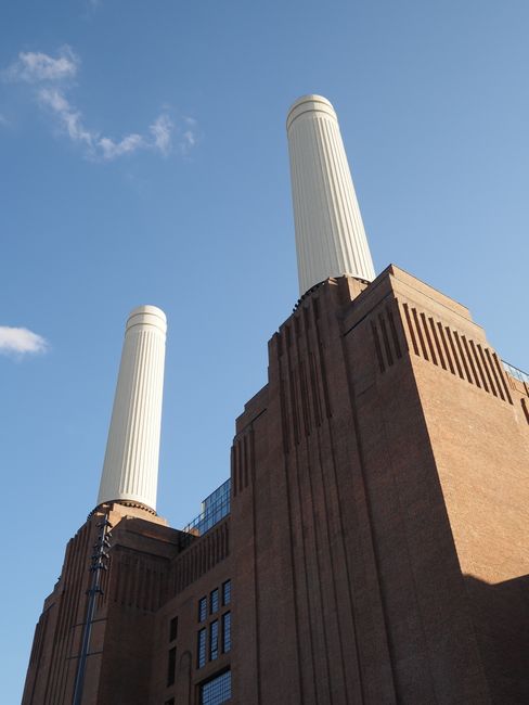 A tall green Christmas tree decorated with gold, silver, and red baubles stands outdoors in front of the Battersea Power Station building, which features a large brick facade with multiple rectangular