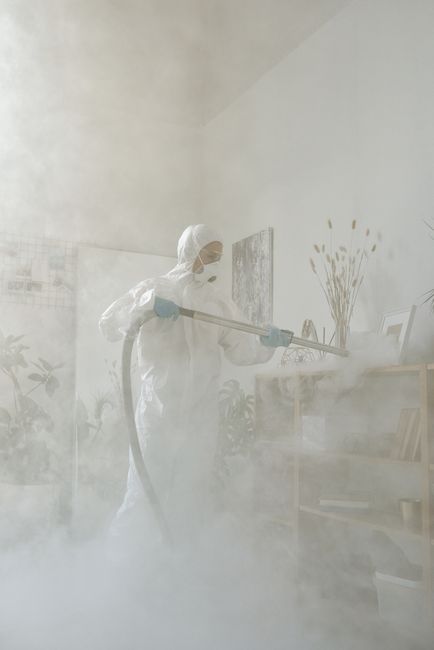 A professional in full protective PPE, including a white suit, mask, and gloves, is engaged in surface cleaning within a residential or commercial indoor space. The individual is holding a high-pressu