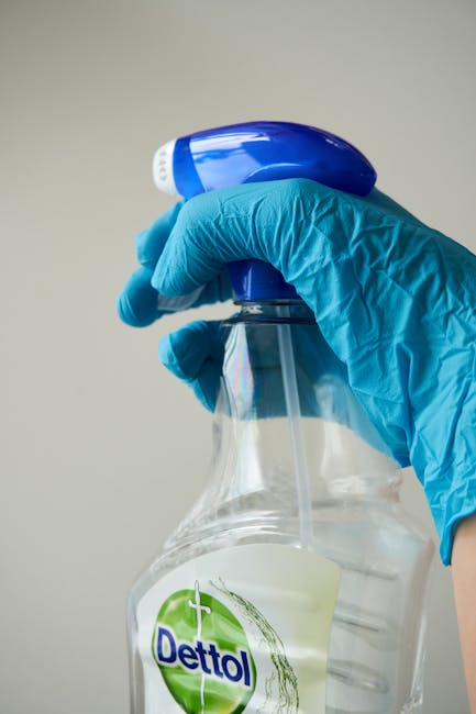 A professional in full protective PPE, including a white suit, mask, and gloves, is engaged in surface cleaning within a residential or commercial indoor space. The individual is holding a high-pressu