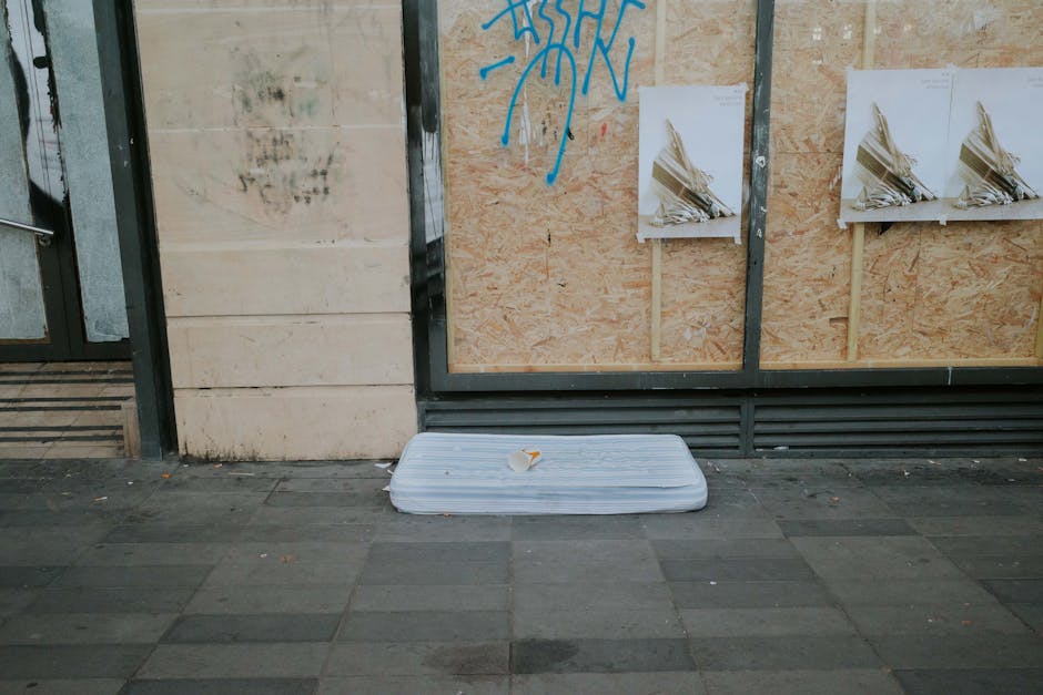 A discarded foam mattress lying flat on a paved sidewalk outside a building with boarded-up windows and graffiti. The mattress appears clean but decommissioned, resting directly on the ground near a c